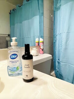 Ein Badezimmer mit einem weißen Waschbecken, einem blauen Duschvorhang und verschiedenen Pflegeprodukten, darunter eine Flasche Dial antibakterielle Defense Handseife, eine Flasche The Well Clean Hands Handdesinfektionsmittel und eine Flasche rosa Lotion.