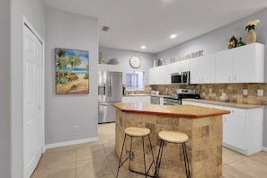 Een keuken met een centraal eiland, barstoelen, een koelkast, magnetron en een klok aan de muur.