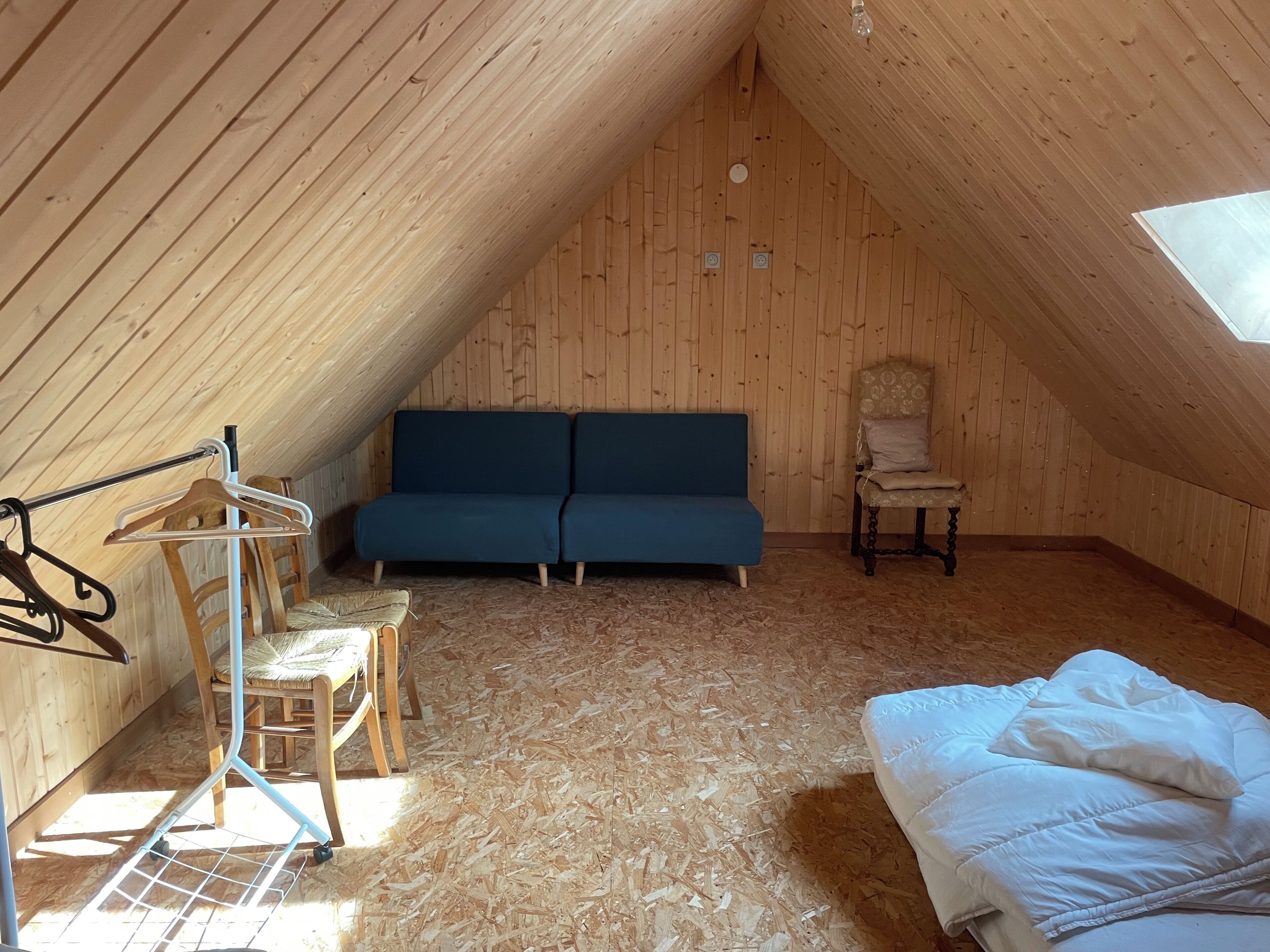 Kodikas ullakkohuone, jossa on puinen, kalteva katto, sininen sohva, puinen tuoli, valkoisilla vuodevaatteilla varustettu sänky ja vaateteline.