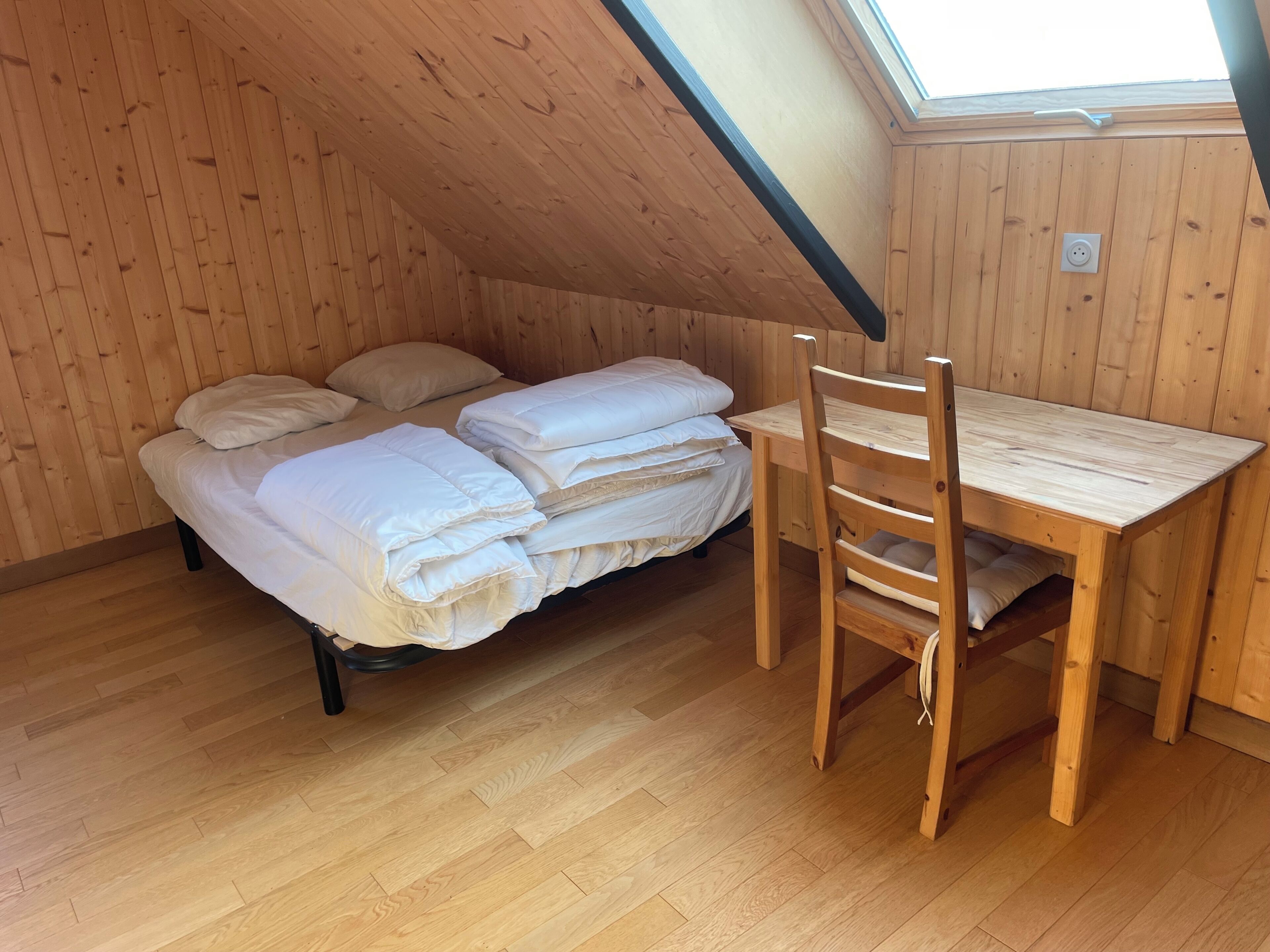 Une petite pièce chaleureuse avec un plafond en pente, des murs en bois, un lit avec une literie blanche, une table en bois et une chaise.