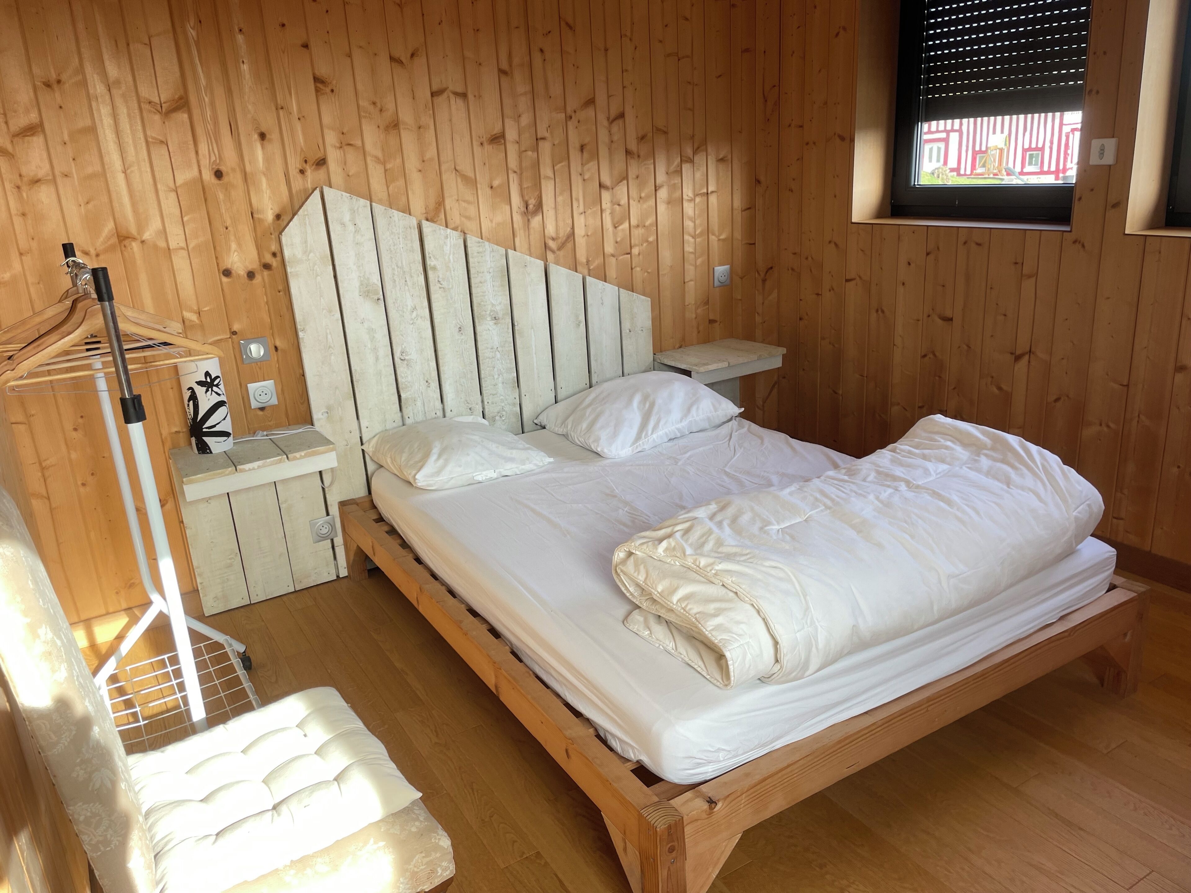 Een kamer met houten lambrisering, een bed, een nachtkastje, een bankje en een raam met jaloezieën.