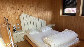 Een kamer met houten lambrisering, een bed, een nachtkastje, een bankje en een raam met jaloezieën.
