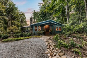 A house with a stone chimney, surrounded by dense forest, and a gravel driveway leading to the entrance.