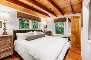 A bedroom with a bed, wooden headboard, bedside tables, lamps, and windows with views of greenery. - New Listing! Mountain Views - Fire pit - Walk to downtown Cashiers (Cashiers)