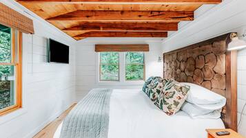 A bedroom with a wooden ceiling, a bed with floral pillows, a bedside table with a lamp, and a window with a view of greenery.