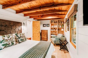 A bedroom with a wooden ceiling, a bed with floral bedding, a wooden wardrobe, and a window with blinds.