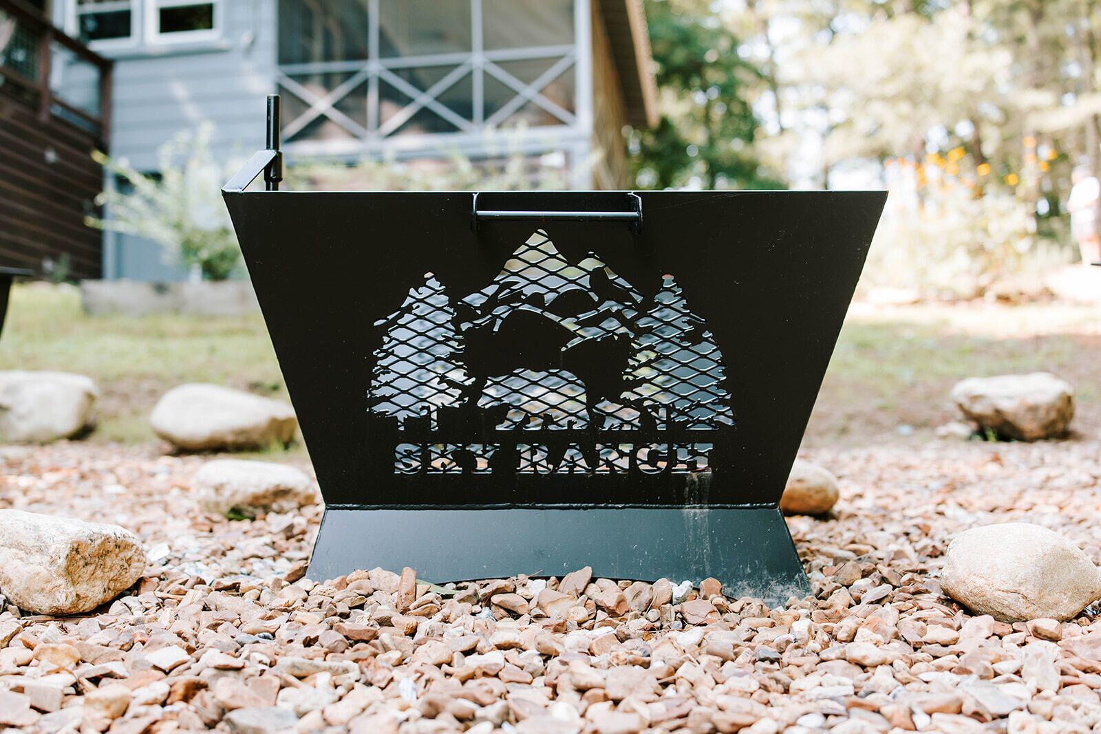 A black metal sign with a mountain and tree silhouette, labeled 'SKY RANCH,' set on a gravel surface with rocks around.