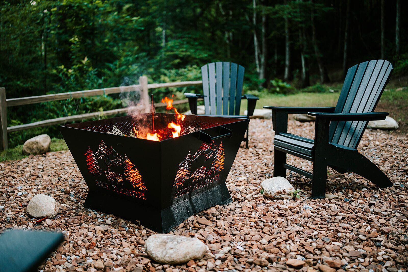 Un foyer avec une grille en métal, entouré de galets, et deux chaises Adirondack à proximité.