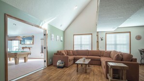 Un salón amplio con un sofá seccional marrón grande, una mesa de centro de madera y un ventanal que da a un área de comedor con mesa de billar.