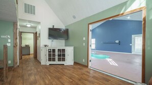Una habitación amplia con piso de madera, un armario blanco, un televisor de pantalla plana montado en la pared y una puerta corredera de vidrio que da a un área exterior.