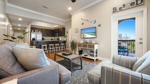 Ein geräumiges Wohnzimmer mit grauem Sofa, Couchtisch und einem gestreiften Sessel. Im Hintergrund ist eine Küche erkennbar, und durch eine Tür ist ein Balkon mit Stadtblick erreichbar.