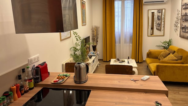 Een moderne woonkamer met een houten salontafel, een gele bank en een keuken met een fornuis en diverse kruiden.