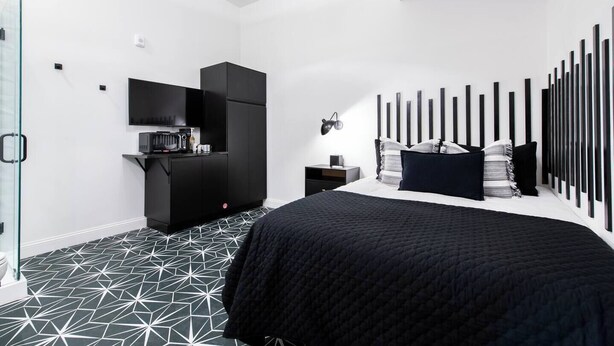 Una camera da letto moderna con pavimento a motivi geometrici, un letto nero con copriletto trapuntato, un'armadio nero, un comodino con una lampada e una TV a muro.