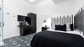 A modern bedroom with a geometric patterned floor, a black bed with a quilted cover, a black wardrobe, a small bedside table with a lamp, and a wall-mounted TV.
