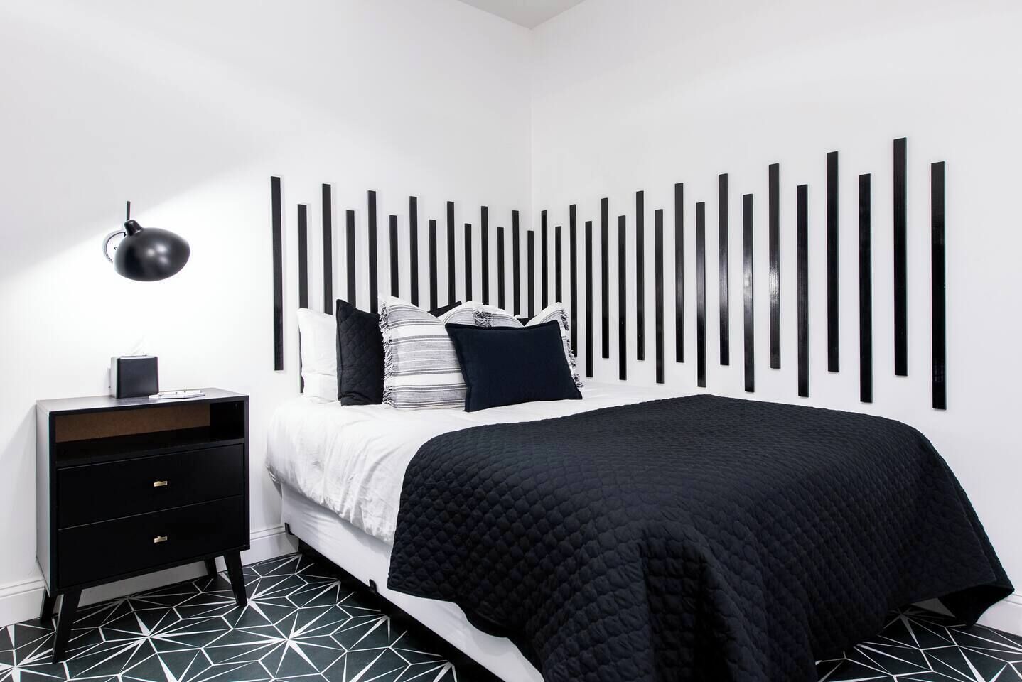 A modern bedroom with a black and white striped headboard, a black bedside table with drawers, and a geometric patterned floor.
