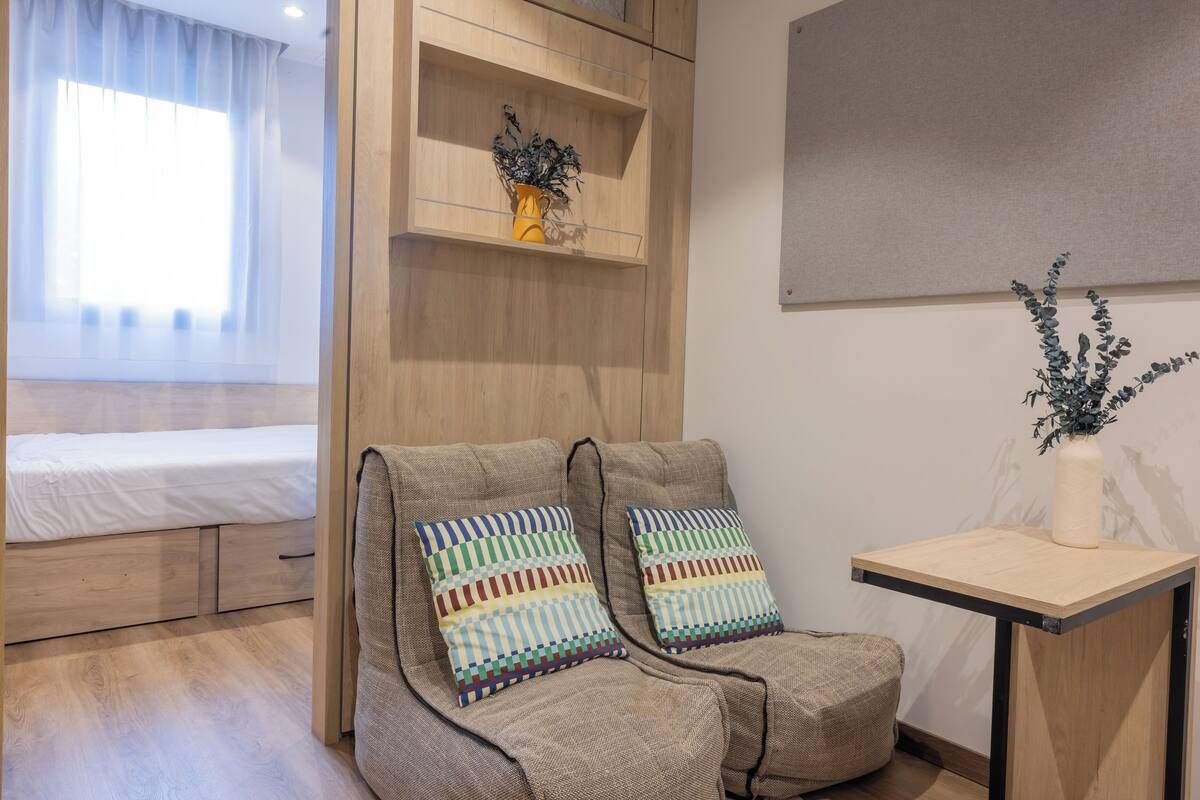 Een moderne kamer met een bed, een sofa met kleurrijke kussens, een tafeltje met een vaas, en een plank met een plant.