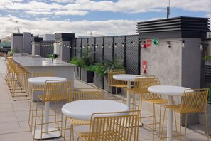 Een buitenruimte met witte ronde tafels en gele stoelen, een grijze muur met een metalen hek en een brandblusser aan de muur bevestigd.