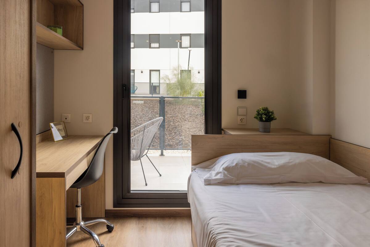 Een moderne slaapkamer met een houten bureau, een comfortabele stoel, een bed met wit beddengoed en uitzicht op het balkon.