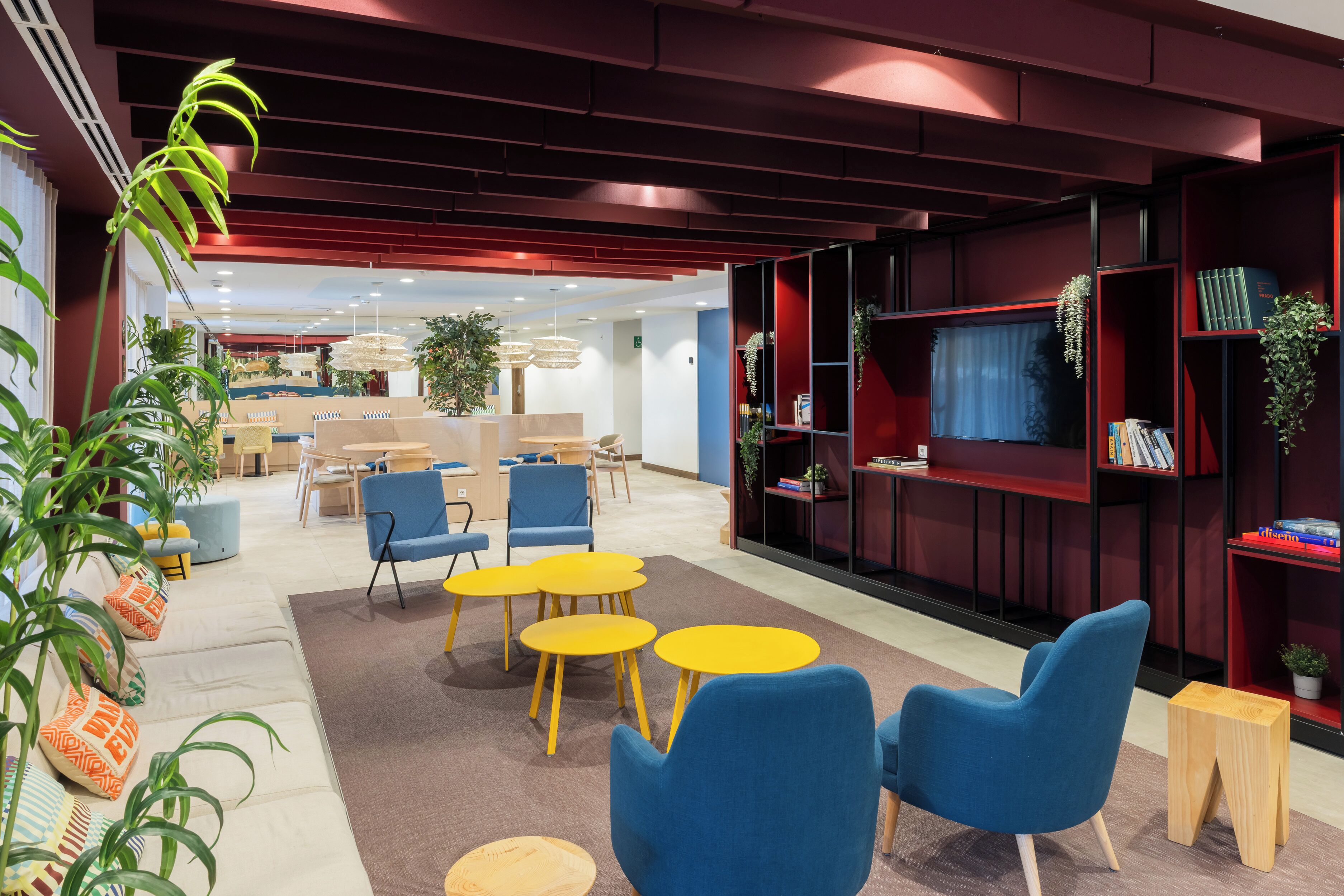 Un área de lounge moderna con sillas azules, mesas amarillas y estanterías rojas.