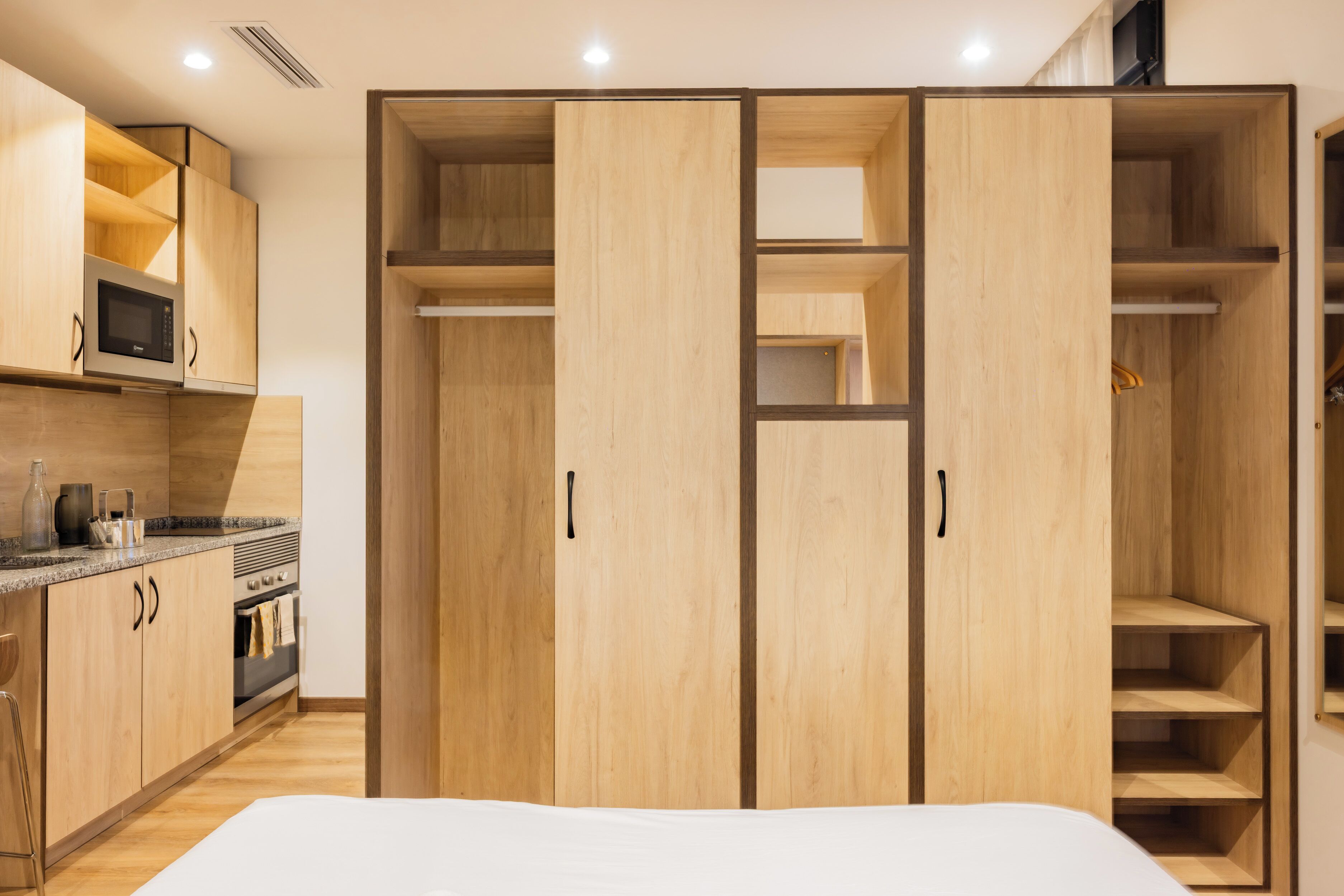 A modern bedroom with a wooden wardrobe, a built-in microwave, and a kitchen area with a stove and cabinets.