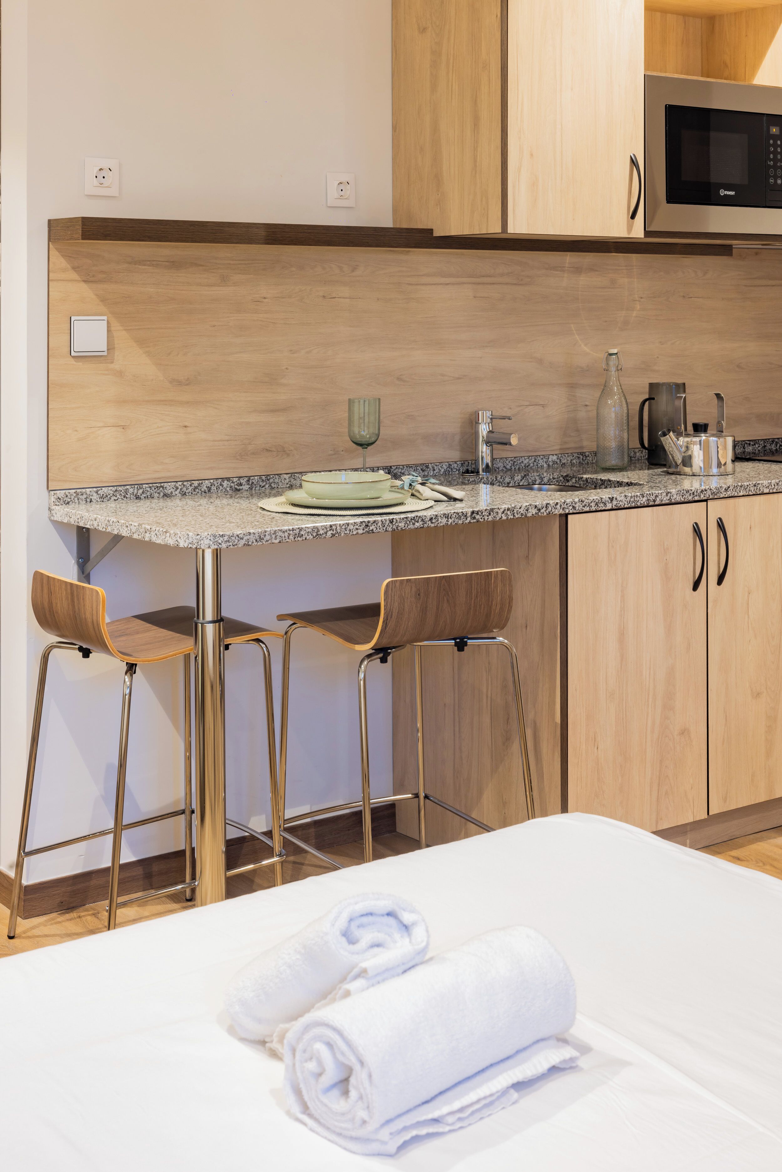 A modern kitchen with wooden cabinets, a granite countertop, and stainless steel appliances. There are two bar stools and a bed with white linens.