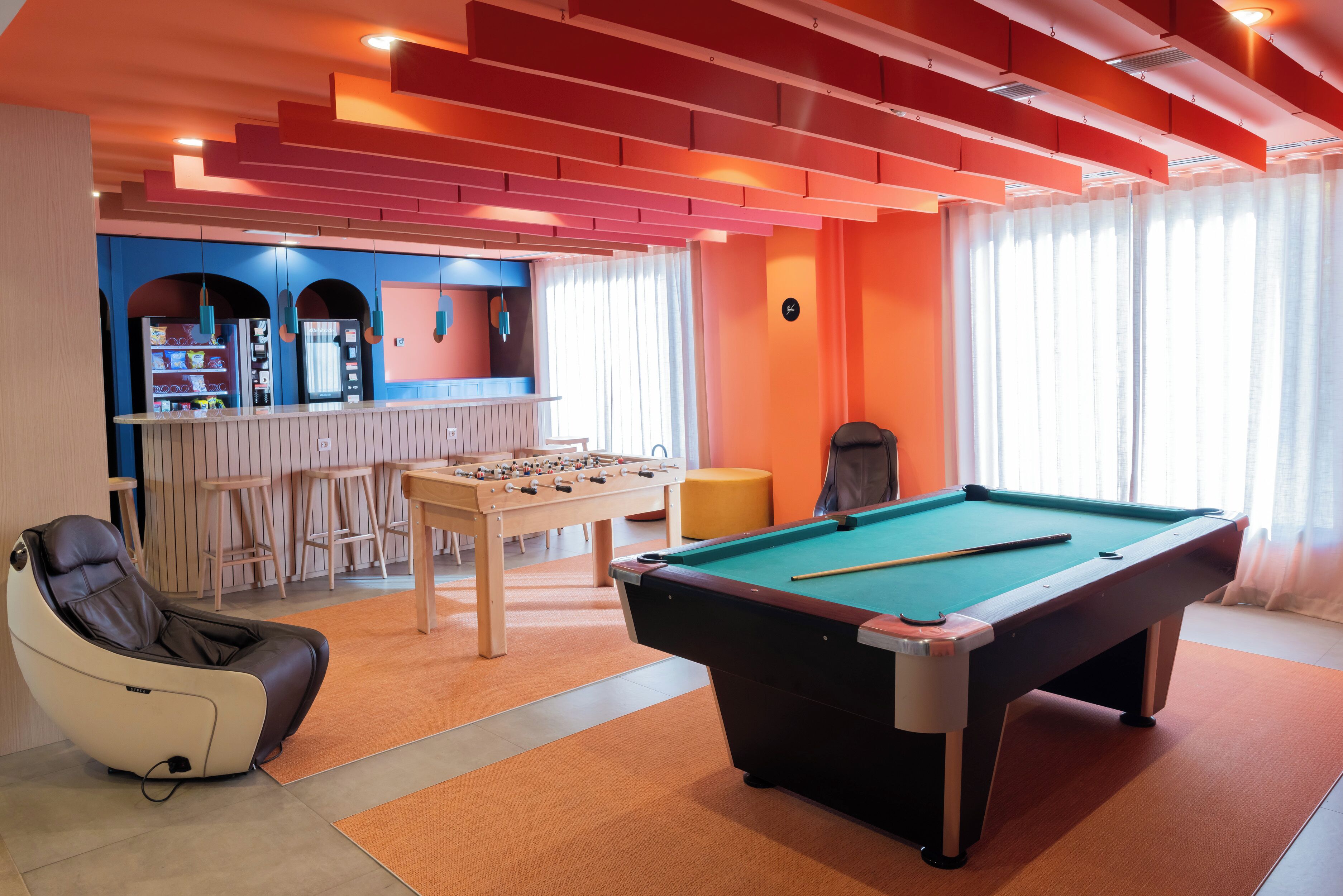 Una mesa de billar, un sillón de masaje y una mesa de futbolín en una habitación con paredes anaranjadas y una zona de bar.