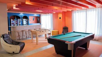 Una mesa de billar, un sillón de masaje y una mesa de futbolín en una habitación con paredes anaranjadas y una zona de bar.