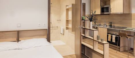 A modern hotel room with a wooden bed, a kitchenette with a microwave and oven, and a shower area.