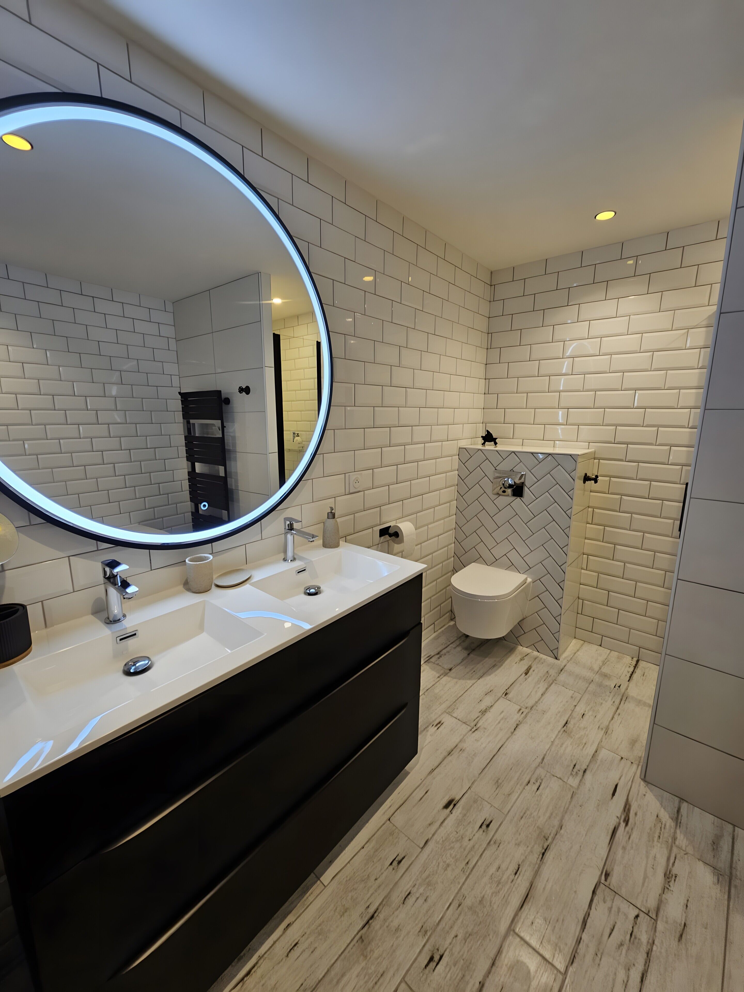 Un baño moderno con azulejos blancos tipo metro, un espejo redondo con iluminación LED, un tocador negro y un recinto de ducha con patrón de espiga.