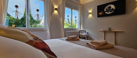 Una habitación de hotel con una cama, dos ventanas con cortinas, una silla, una lámpara de pared y un cartel.