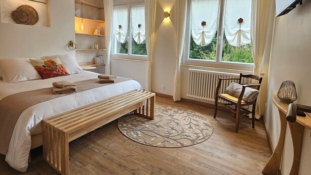 Ein Schlafzimmer mit Bett, Nachttischen, einem Sessel, einem Fenster mit Vorhängen, einer Heizung und einer Decke mit Holzbalken.