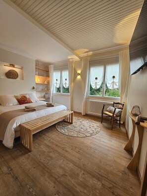 Une chambre à coucher comprenant un lit, des tables de chevet, une chaise, une fenêtre avec des rideaux, un radiateur et un plafond avec des lambris en bois.