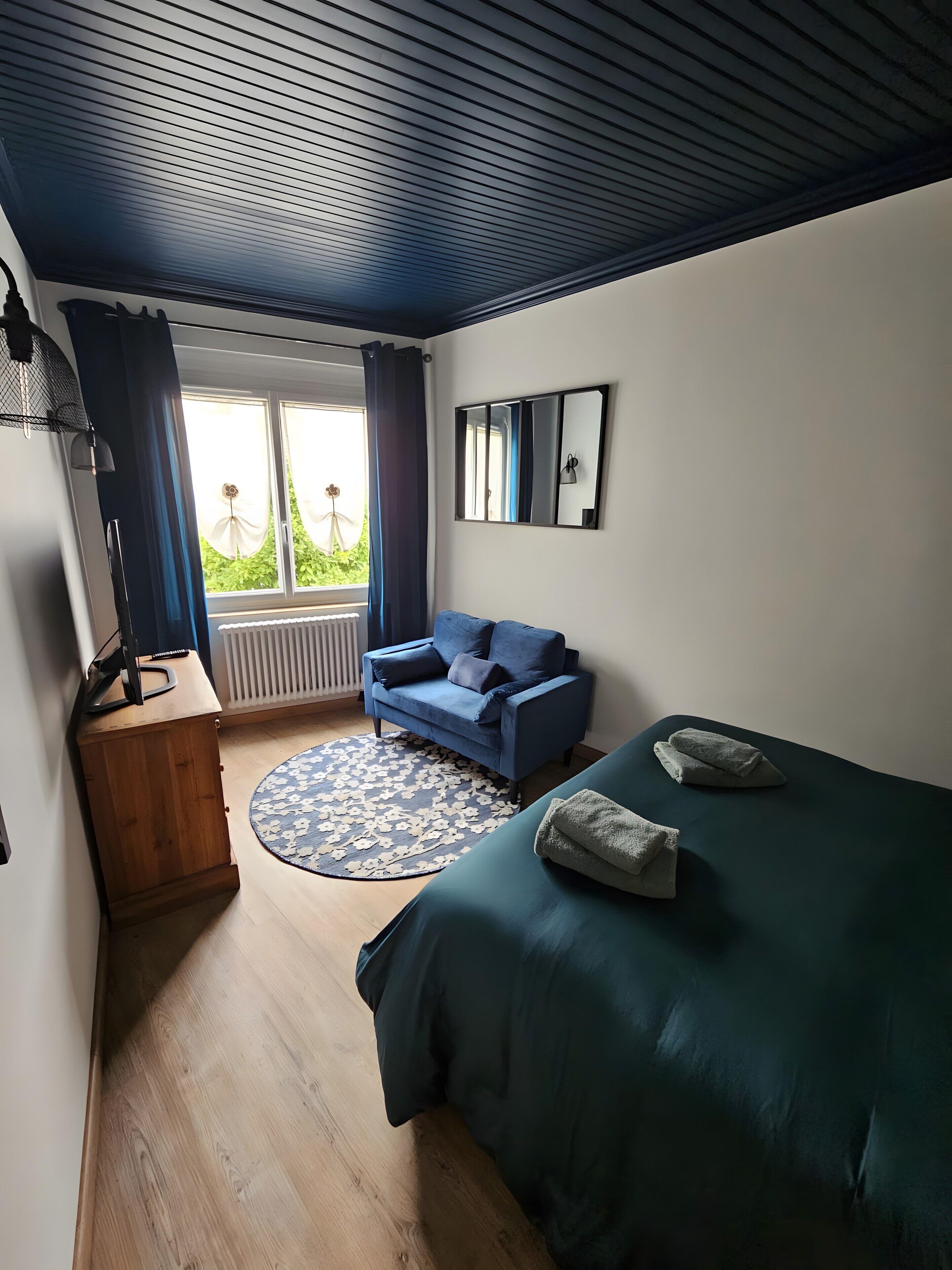 A bedroom with a bed, a blue sofa, a wooden cabinet, a round rug, and a window with curtains.