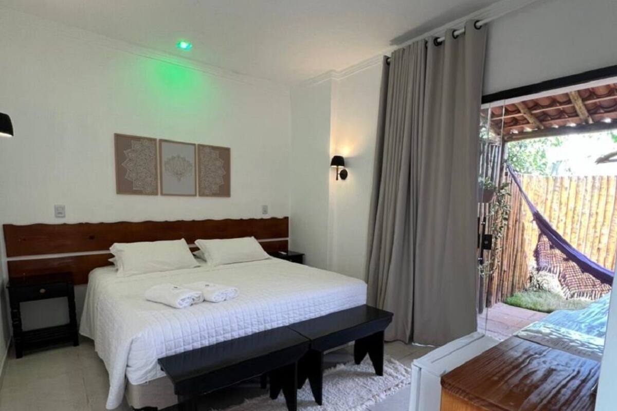 Une chambre à coucher comprenant un lit, une table de chevet, un banc et donnant sur un espace extérieur avec un hamac.