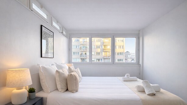 A minimalist bedroom with a large bed, a bedside table with a lamp, and a window with a city view. - Appartement pres du parc Montsouris (Paris)
