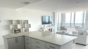 Uma cozinha moderna com armários brancos e bancada de mármore, que se abre para uma sala de estar com sofá branco e televisão na parede.