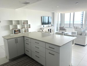 Eine moderne Küche mit weißen Schränken und einer Marmorarbeitsplatte, die in einen Wohnbereich mit einem weißen Sofa und einem an der Wand befestigten Fernseher übergeht.