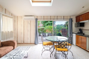 A compact kitchen with a dining area, a sofa, and a view of greenery. - Gîte Navacelles (Lavérune)