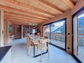 A wooden interior with a dining area, bookshelves, and a large window overlooking a mountainous landscape.