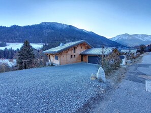 A wooden cabin with a sloped roof, a gravel driveway, and a mountainous backdrop.