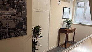 A hallway with a large cityscape painting, a potted plant, a small table with a vase, and a window with blinds.