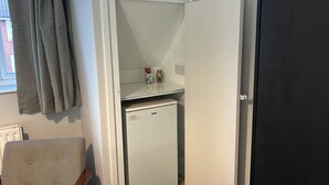 A compact room with a white refrigerator, a small shelf with items, a window with curtains, and a door.