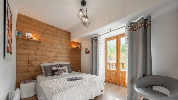 Een slaapkamer met een houten hoofdbord, een bed met een witte deken, een grijze stoel en een raam met gordijnen.