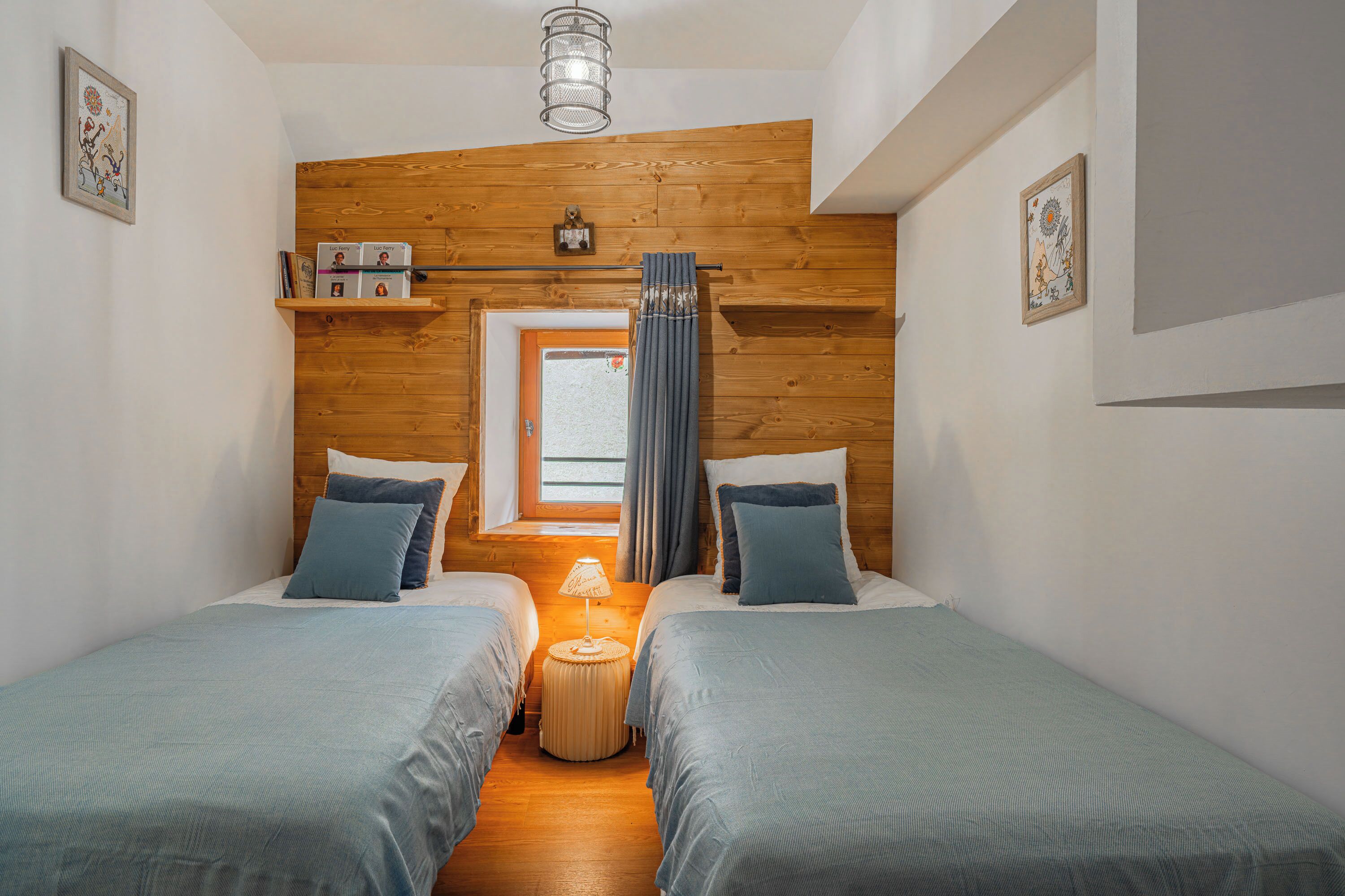 Een kamer met twee bedden, een klein rond tafeltje, een raam met gordijnen, houten lambrisering en een plafondlamp.