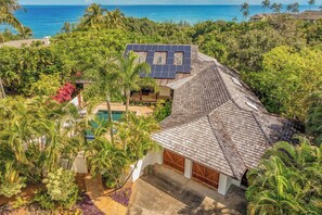 A house with a sloped roof, solar panels, and a swimming pool surrounded by tropical vegetation.
