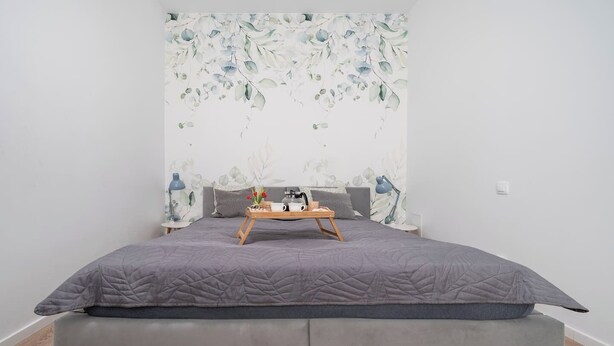 Een moderne slaapkamer met een grijs gewatteerd bed, een houten nachtkastje met een dienblad waarop een kopje en een boek staan, en een muur met bloemenbehang.