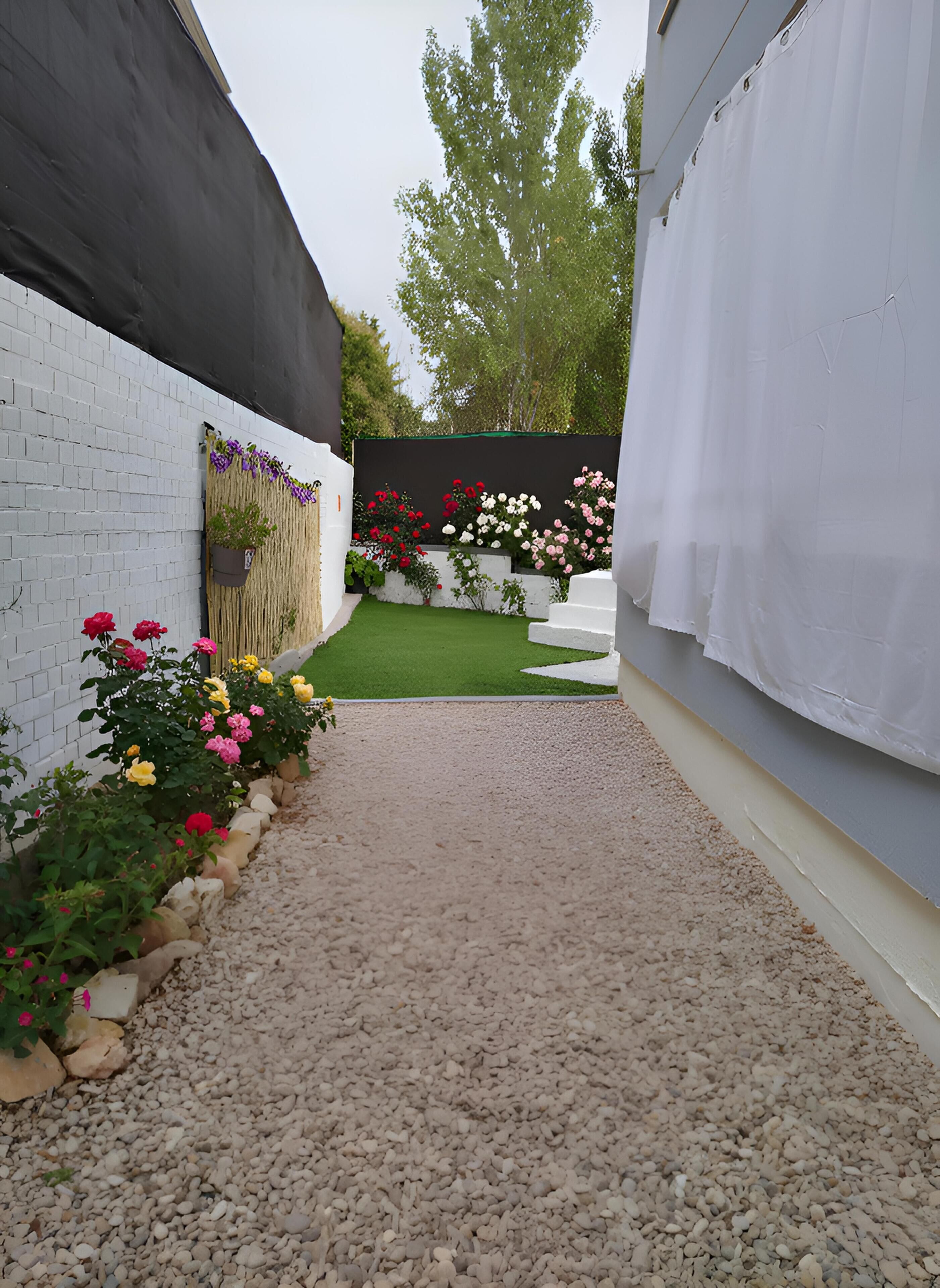 Un estrecho sendero de grava que conduce a un jardín con un muro de ladrillo blanco, un toldo negro y una cerca blanca.