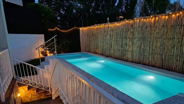 A swimming pool with a bamboo wall and string lights, surrounded by a white fence and stairs leading down to the pool. - YOUR OASIS NEAR MADRID WITH A HOT TUB AND SWIMMING POOL. (Villarejo de Salvanés)