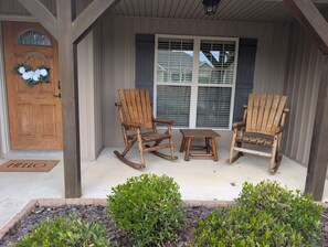 A porch with two rocking chairs and a small table, a wooden door with a wreath, and a window with blinds.