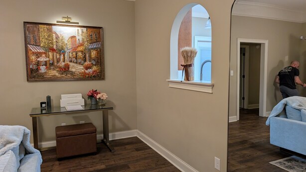 Una habitación con piso de madera, una mesa de vidrio con un florero y flores, un cuadro en la pared y una ventana con cortina.